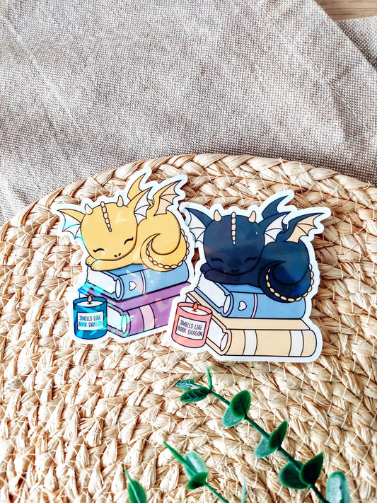 Two holographic vinyl stickers featuring sleeping baby dragons resting on stacked books with small candles, photographed on a woven neutral background.