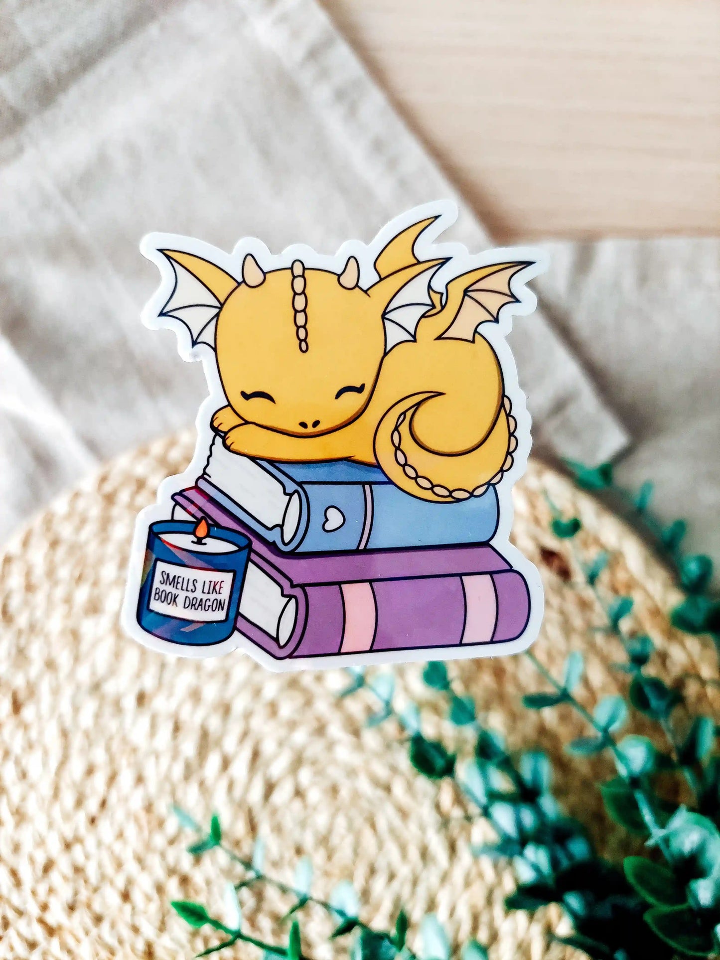 Holographic vinyl sticker of a golden baby dragon sleeping on stacked books with a candle.