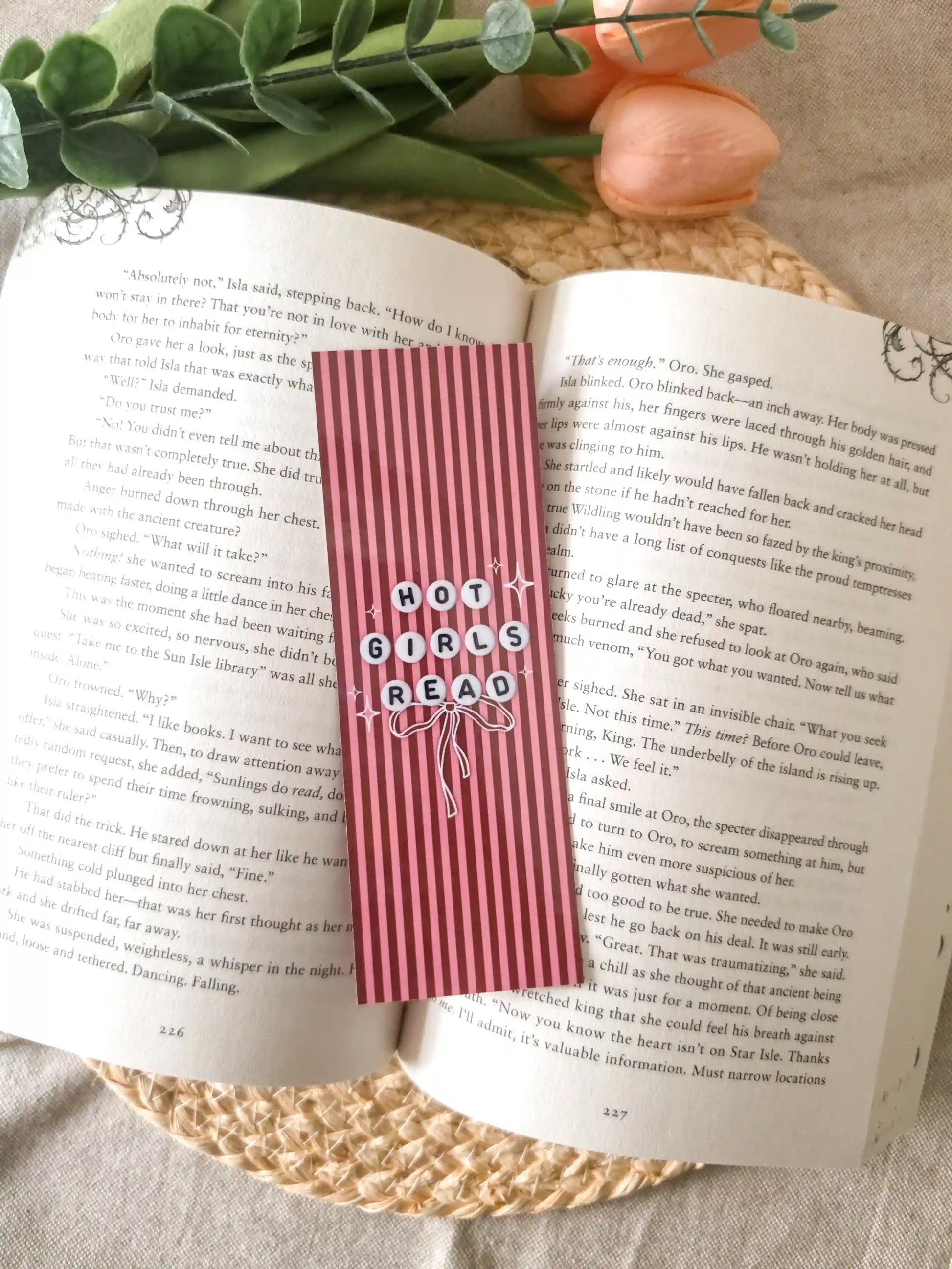 A pink striped book bookmark with the words 'HOT GIRLS READ' in white lettering style like friendship bracelets beads, placed on an open book with decorative greenery and fruit around it.