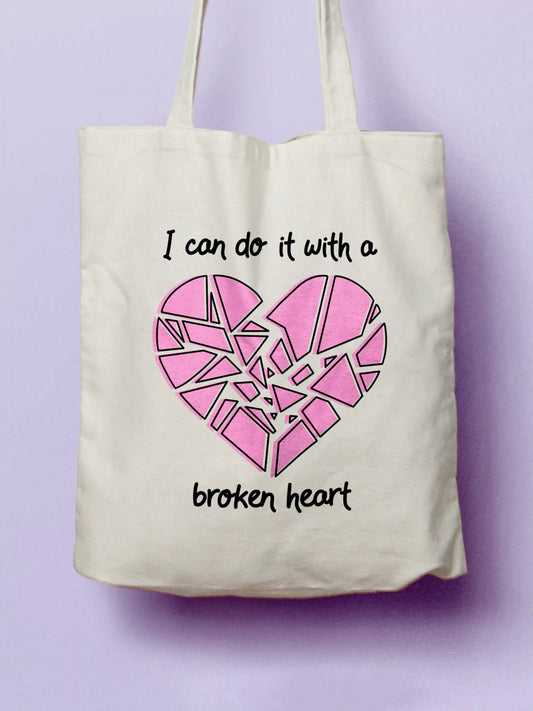 Cotton canvas tote bag with a pink broken heart graphic and the text “I can do it with a broken heart,” photographed on a soft purple background.