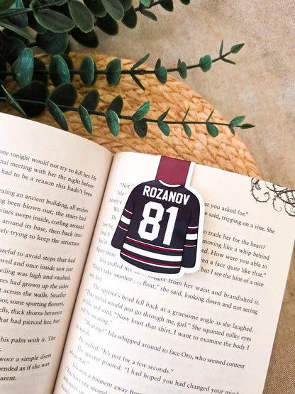 Heated Rivalry inspired hockey jersey magnetic bookmark clipped onto an open book, showing the blue and red jersey design with "Rozanov 81" on the back.