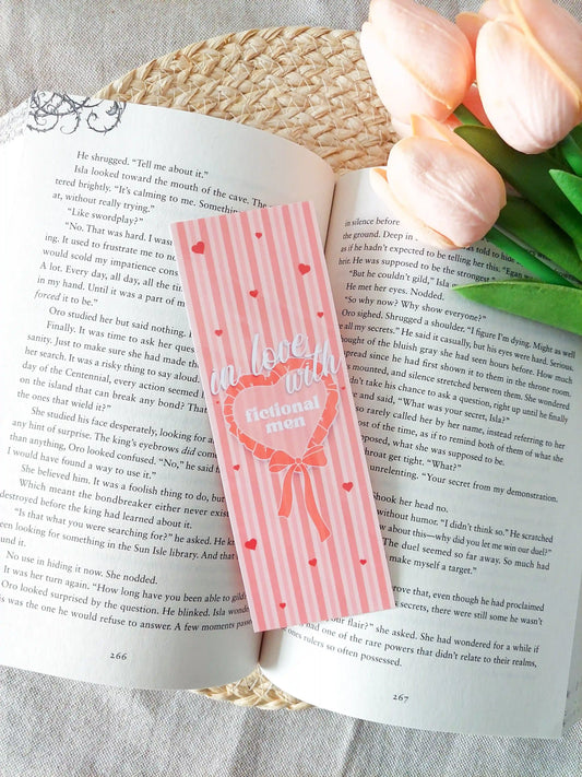 Bookmark with striped pink and peach background, small heart details and bow illustration, featuring the text "In love with fictional men", placed on an open book.