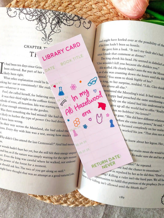 Bookmark designed like a library card, with the text “In my Ali Hazelwood era”, and small themed illustrations, resting on an open book.