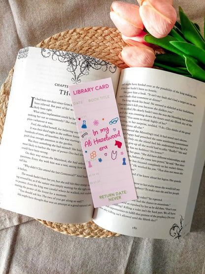 Bookmark designed like a library card, with the text “In my Ali Hazelwood era”, and small themed illustrations, resting on an open book.