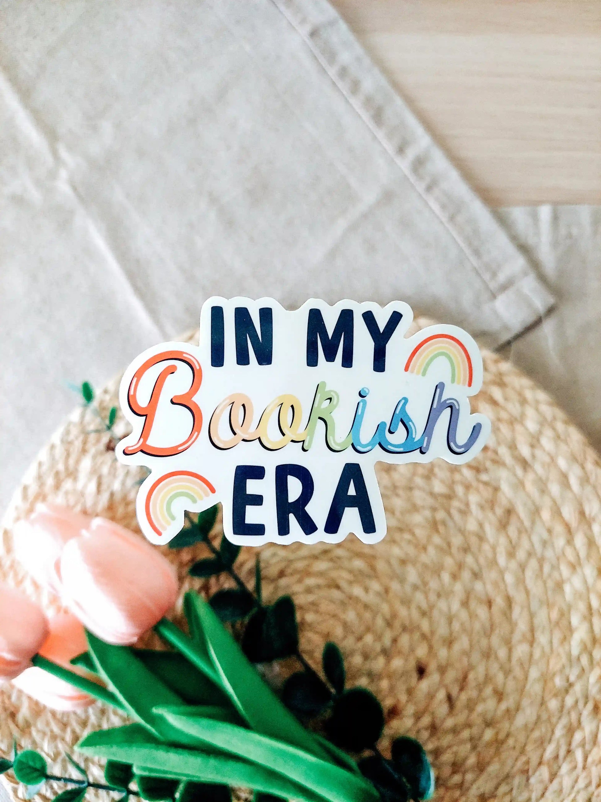 Vinyl sticker featuring the text "In My Bookish Era" in colorful rainbow lettering and small rainbow illustrations, photographed on neutral fabric background with woven mat.