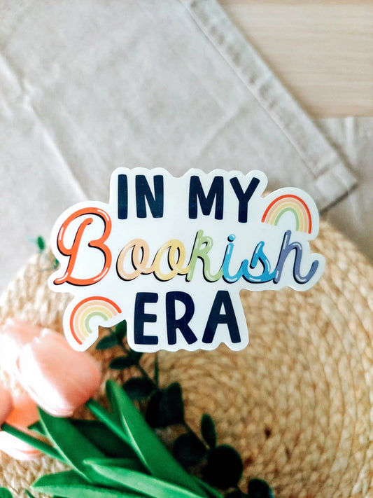 Vinyl sticker featuring the text "In My Bookish Era" in colorful rainbow lettering and small rainbow illustrations, photographed on neutral fabric background with woven mat.