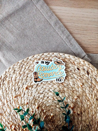Vinyl sticker featuring the text "In My Cowboy Romance Era" in western lettering with cowboy boot, cowboy hat, and heart illustrations, photographed on neutral fabric background with woven mat.