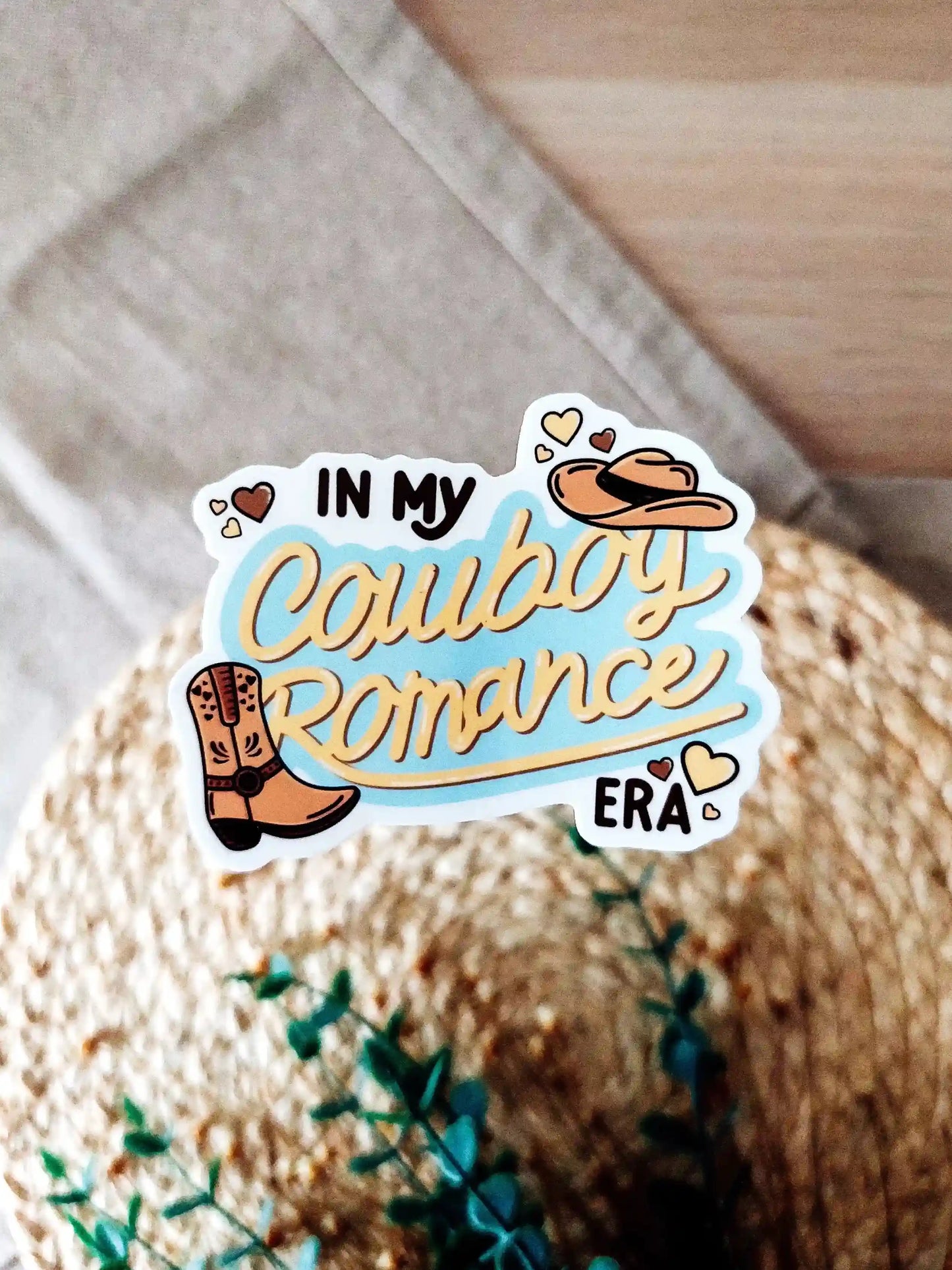 Vinyl sticker featuring the text "In My Cowboy Romance Era" in western lettering with cowboy boot, cowboy hat, and heart illustrations, photographed on neutral fabric background with woven mat.