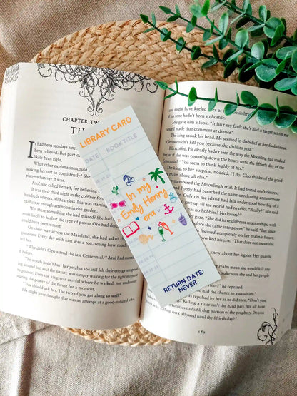 Bookmark designed like a library card, with the text “In my Emily Henry era”, and small themed illustrations, resting on an open book.