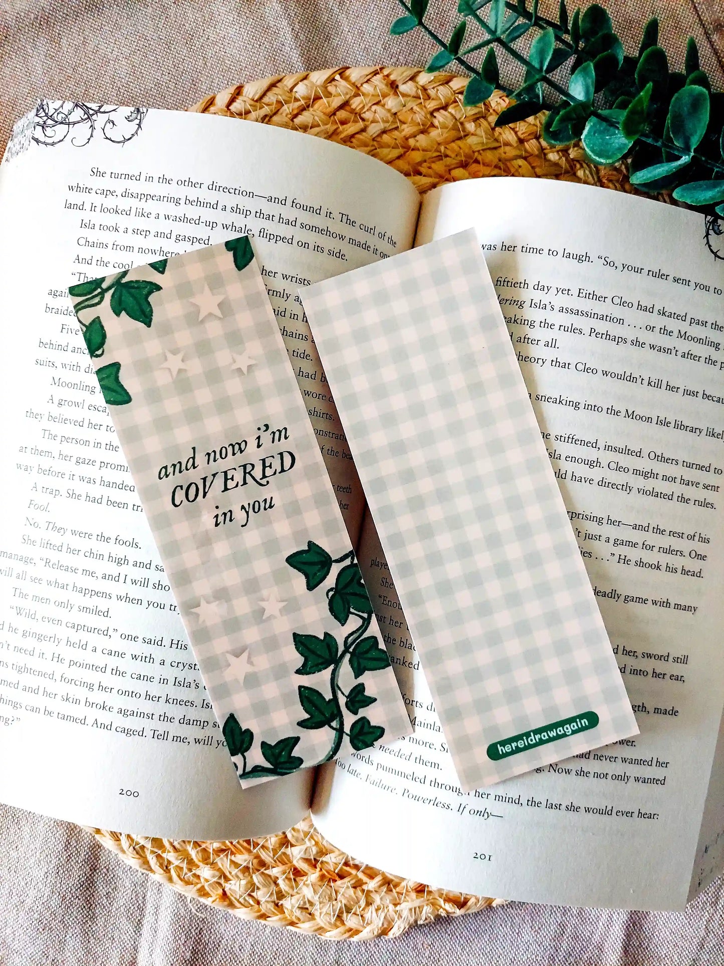 Bookmark inspired by Taylor Swift's Ivy featuring ivy illustrations and green gingham background, with the lyric "and now I'm covered in you", displayed on an open book over a woven mat surface decorative plants.