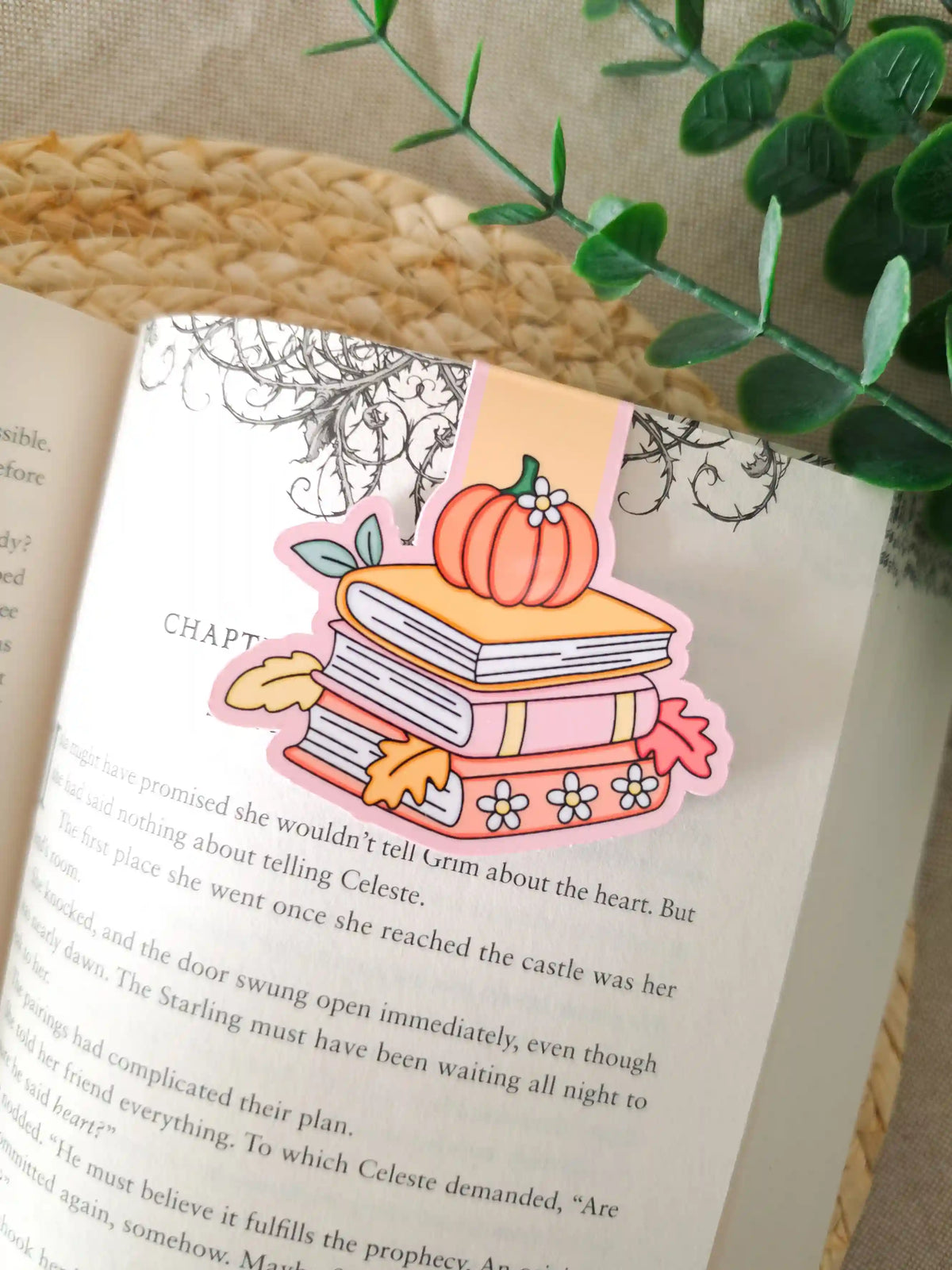 A die-cut magnetic bookmark featuring an illustrated stack of books in pink and yellow with a small orange pumpkin and white daisies on top, surrounded by yellow and red autumn leaves, clipped onto the top corner of an open book resting on a woven rattan mat, with green eucalyptus sprigs in the upper right corner.
