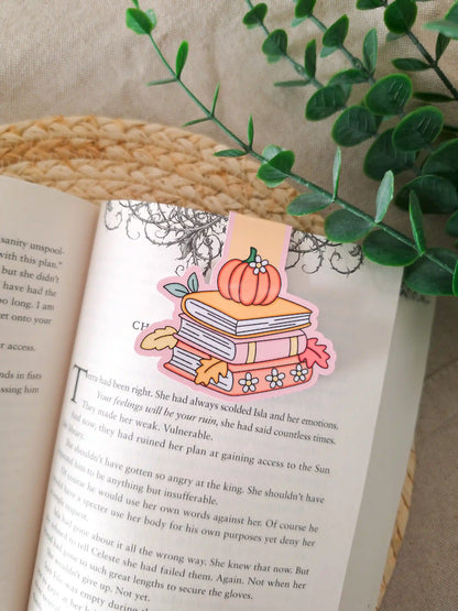 A die-cut magnetic bookmark featuring an illustrated stack of books in pink and yellow with a small orange pumpkin and white daisies on top, surrounded by yellow and red autumn leaves, clipped onto the top corner of an open book resting on a woven rattan mat, with green eucalyptus sprigs in the upper right corner.