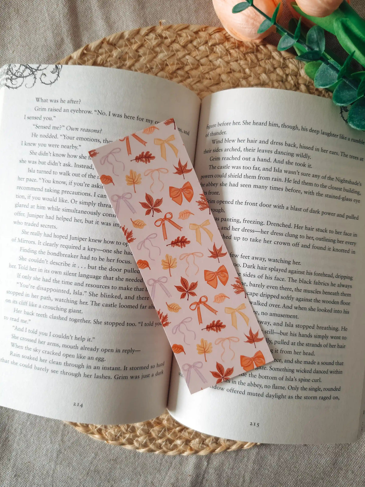 A laminated bookmark featuring scattered illustrated autumn leaves in shades of orange, red, and brown, and decorative bows in orange, yellow, and lavender on a pale blush background, resting on an open book on a woven rattan mat, with green foliage visible in the upper right corner.