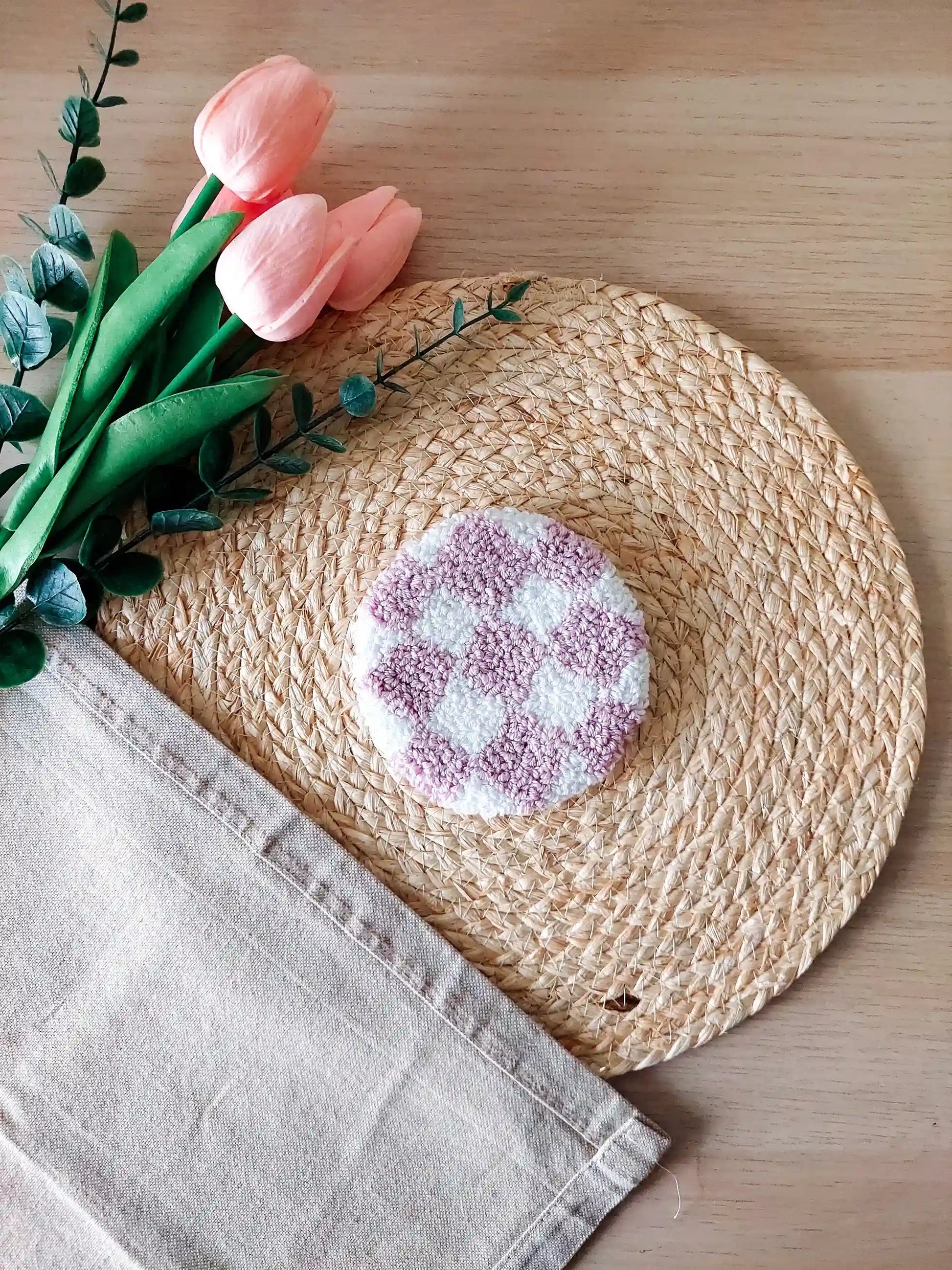 Handmade round punch needle coaster featuring a lilac and white checkered pattern, photographed on a woven mat with pink decorative flowers in the background.