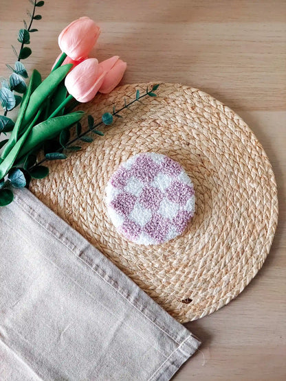 Handmade round punch needle coaster featuring a lilac and white checkered pattern, photographed on a woven mat with pink decorative flowers in the background.