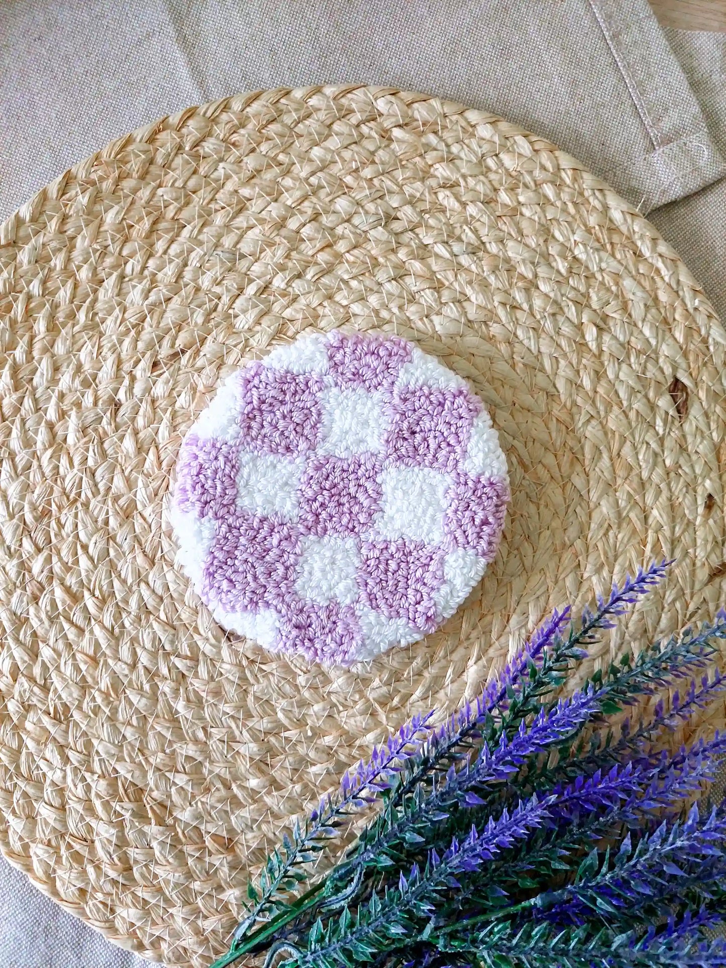 Handmade round punch needle coaster featuring a lilac and white checkered pattern, photographed on a woven mat with purple decorative flowers in the background.