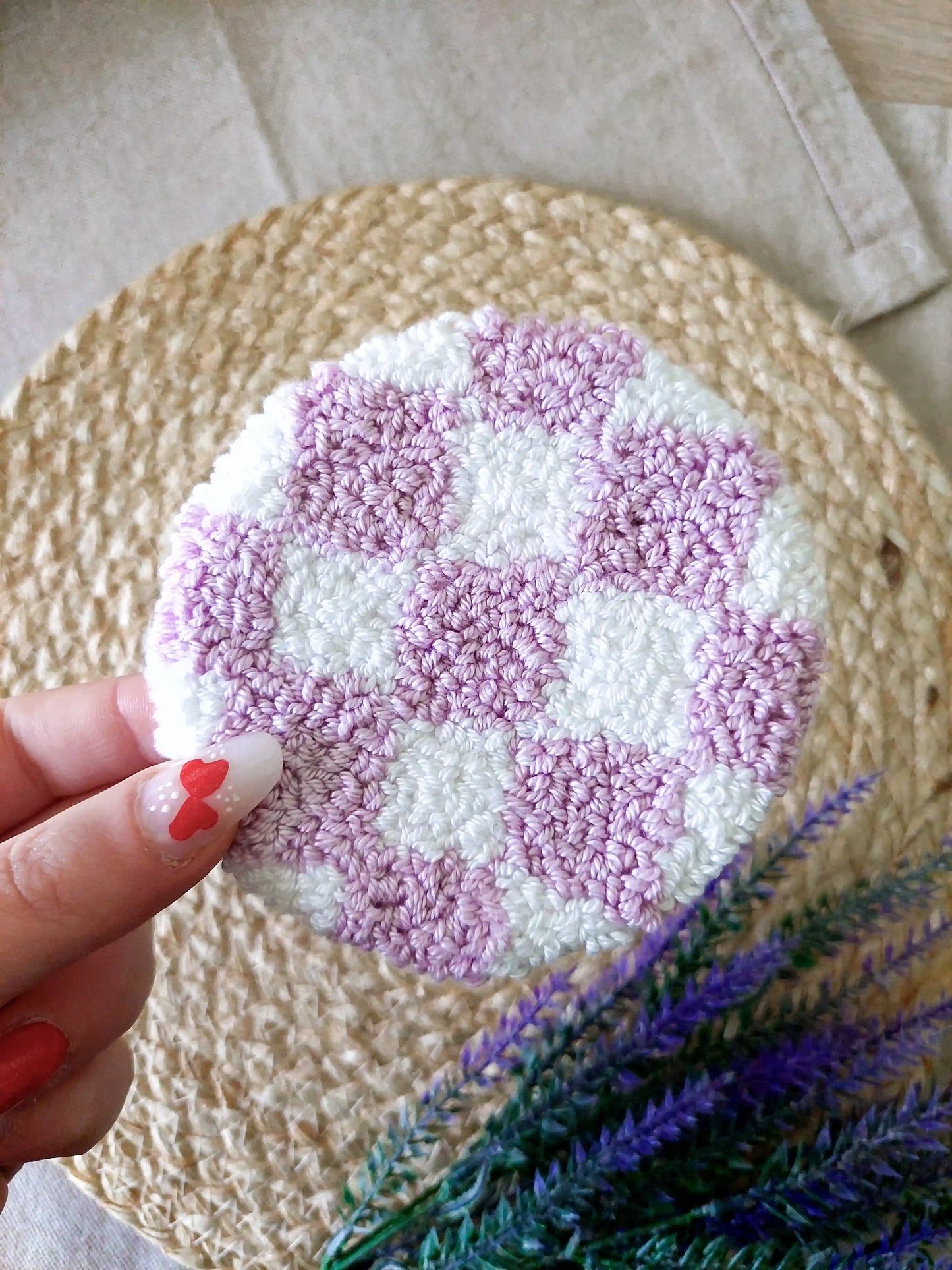 Handmade round punch needle coaster featuring a lilac and white checkered pattern, photographed on a woven mat with purple decorative flowers in the background.