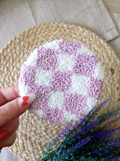 Handmade round punch needle coaster featuring a lilac and white checkered pattern, photographed on a woven mat with purple decorative flowers in the background.