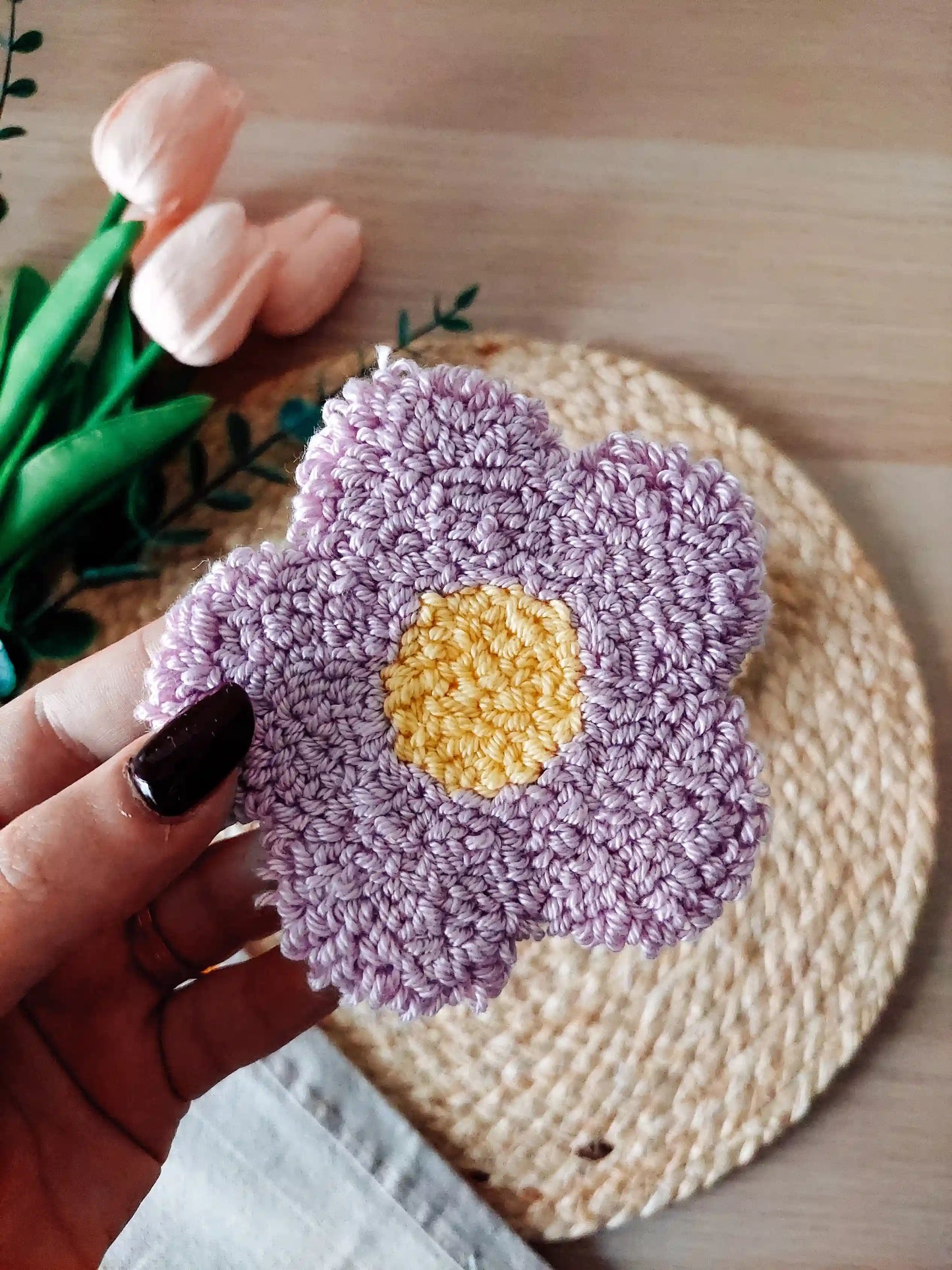 Handmade punch needle coaster shaped like a lilac daisy flower with a yellow center, photographed on a woven mat with decorative flowers in the back.