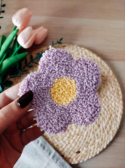Handmade punch needle coaster shaped like a lilac daisy flower with a yellow center, photographed on a woven mat with decorative flowers in the back.