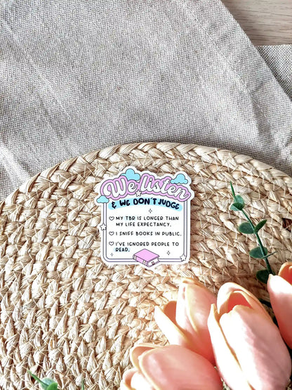 Glossy vinyl sticker with pastel pink and blue design reading "We listen & we don’t judge" followed by funny book lover confessions: "My TBR is longer than my life expectancy. I sniff books in public. I’ve ignored people to read". Photographed on a neutral fabric background with woven mat and pink tulips.