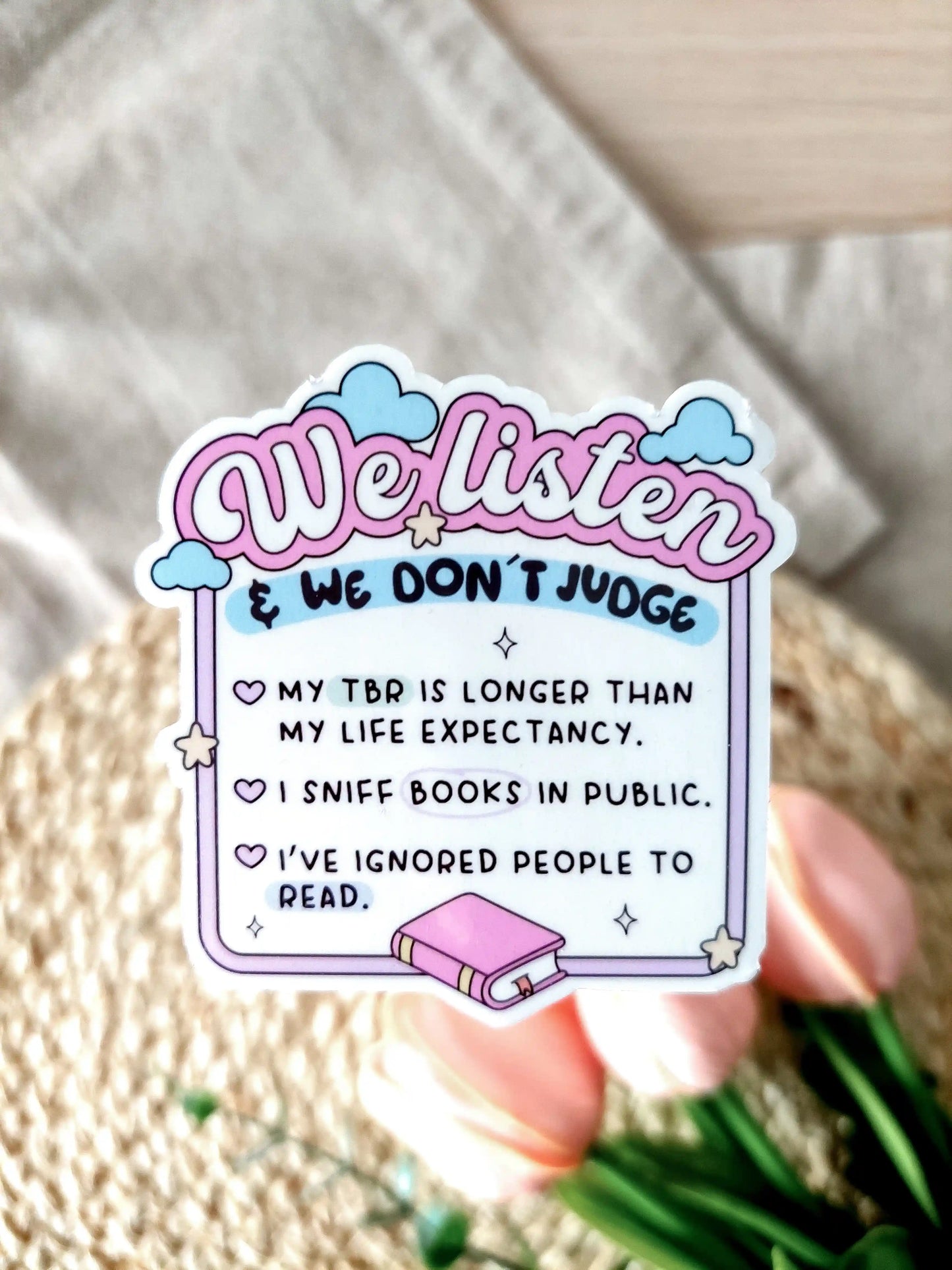 Glossy vinyl sticker with pastel pink and blue design reading "We listen & we don’t judge" followed by funny book lover confessions: "My TBR is longer than my life expectancy. I sniff books in public. I’ve ignored people to read". Photographed on a neutral fabric background with woven mat and pink tulips.