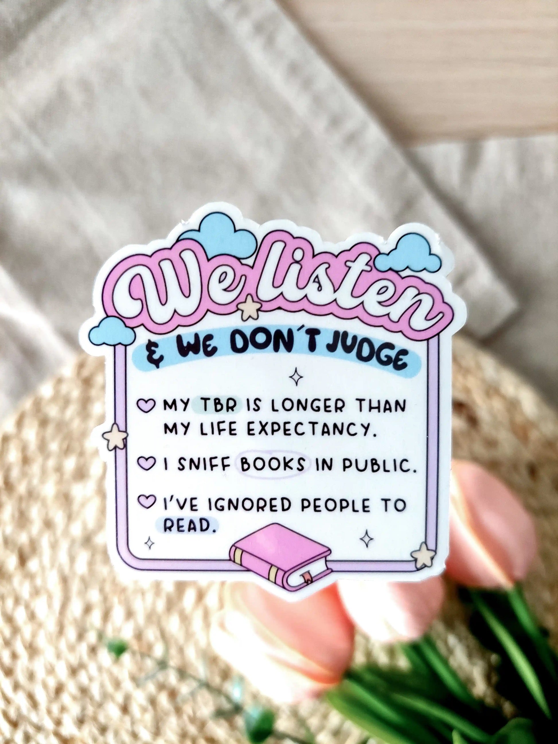 Glossy vinyl sticker with pastel pink and blue design reading "We listen & we don’t judge" followed by funny book lover confessions: "My TBR is longer than my life expectancy. I sniff books in public. I’ve ignored people to read". Photographed on a neutral fabric background with woven mat and pink tulips.