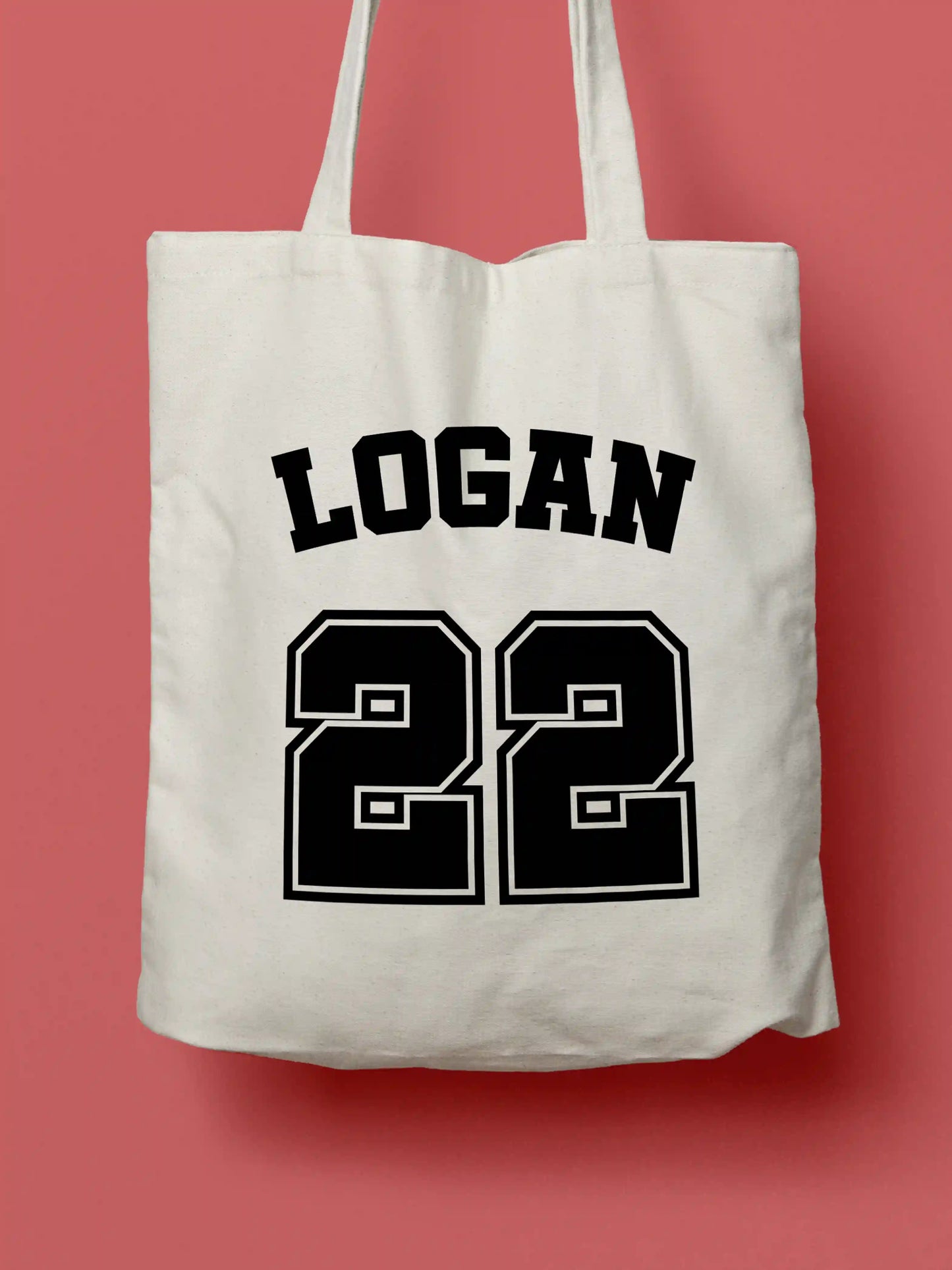 A white canvas tote bag featuring "Logan 22" in bold black collegiate-style lettering, photographed against a lavender background.