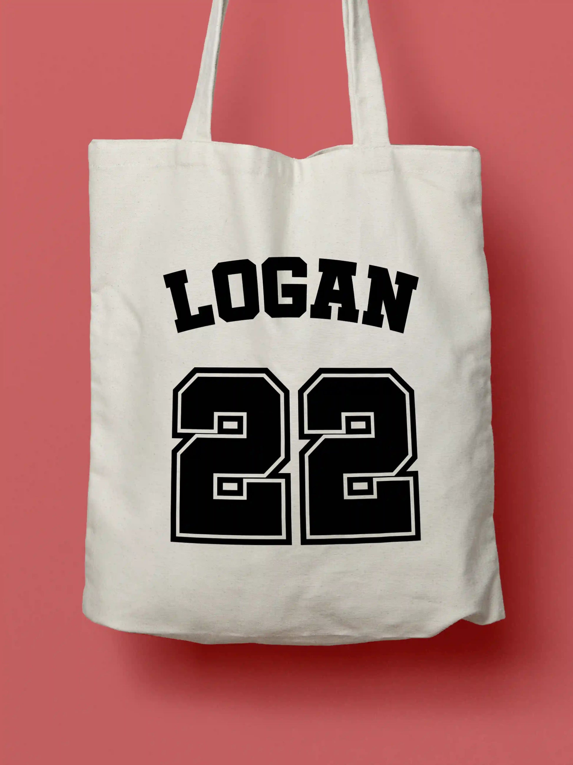 A white canvas tote bag featuring "Logan 22" in bold black collegiate-style lettering, photographed against a lavender background.