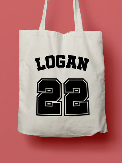 A white canvas tote bag featuring "Logan 22" in bold black collegiate-style lettering, photographed against a lavender background.