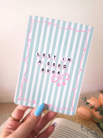A hand holding an e-reader insert with a light blue and white striped background, a pink decorative border, the text "Lost in a Good Book" in letter bead-style lettering, and a pink bow with cherry charms detail, placed on an open book resting on a woven rattan mat. 