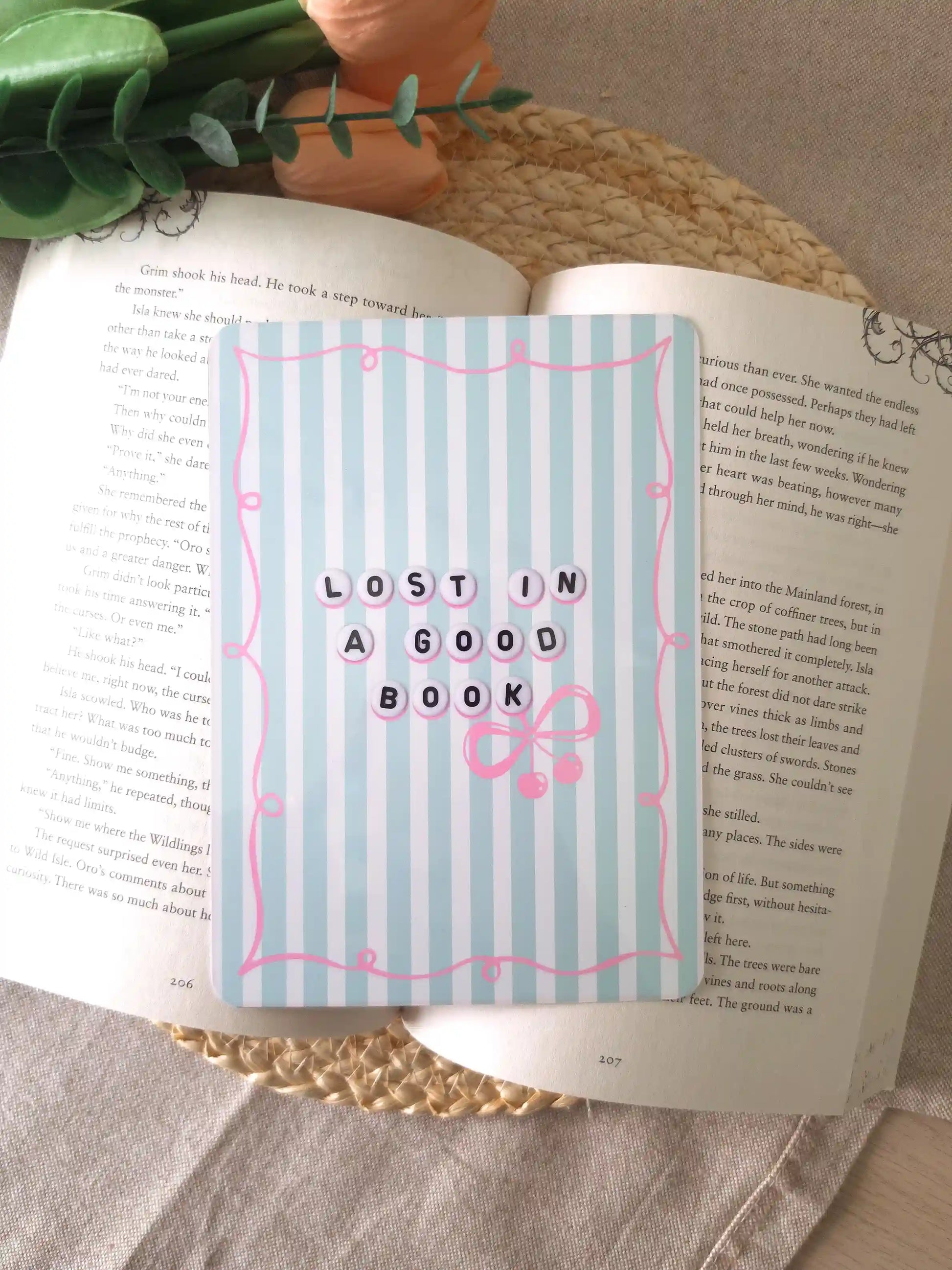An e-reader insert with a light blue and white striped background, a pink decorative border, the text "Lost in a Good Book" in letter bead-style lettering, and a pink bow with cherry charms detail, placed on an open book resting on a woven rattan mat. 