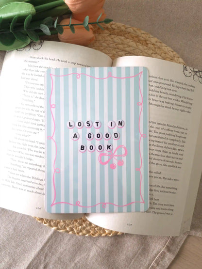 An e-reader insert with a light blue and white striped background, a pink decorative border, the text "Lost in a Good Book" in letter bead-style lettering, and a pink bow with cherry charms detail, placed on an open book resting on a woven rattan mat. 