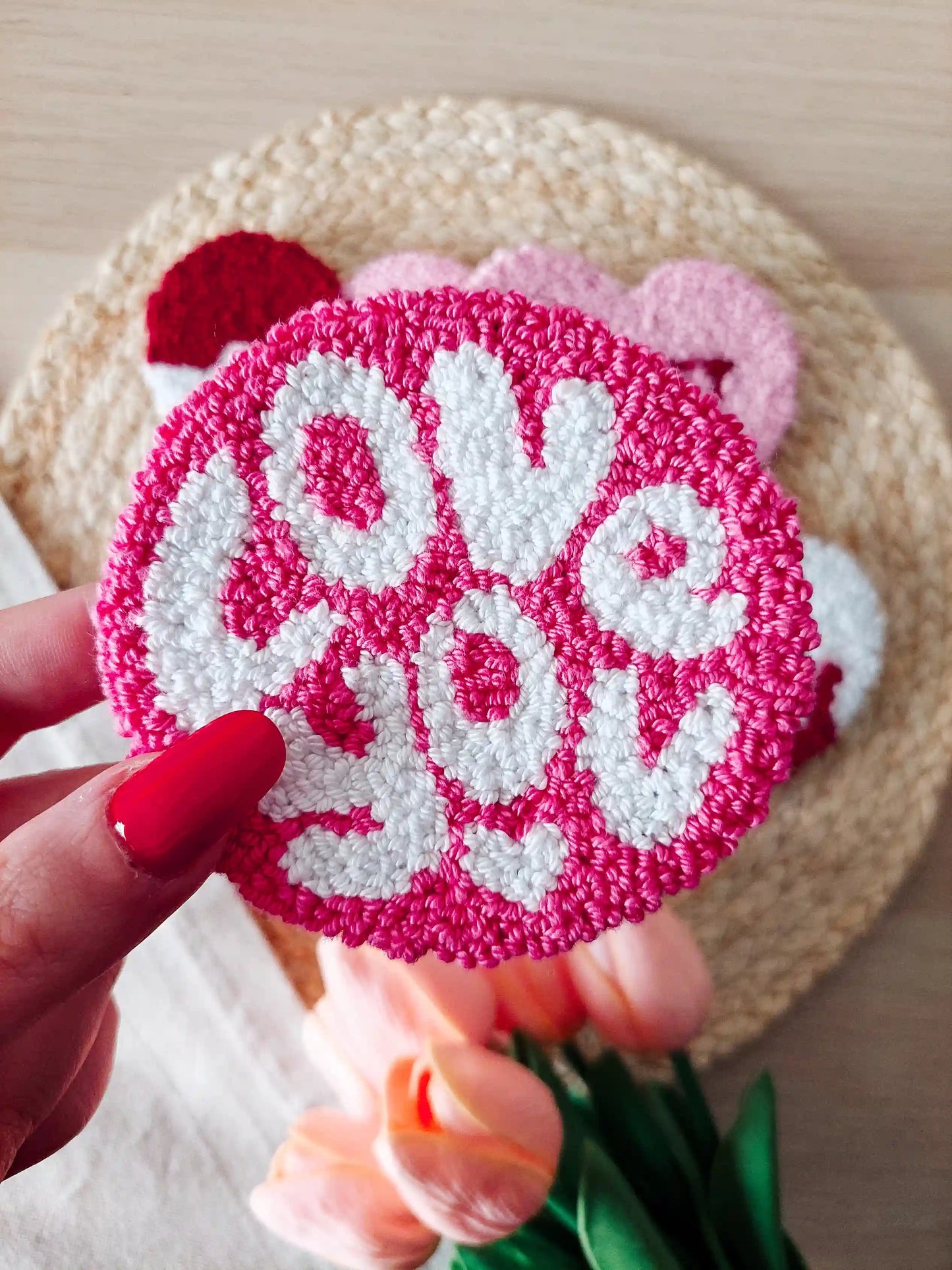 Handmade pink punch needle coaster with white "Love You" lettering, displayed on a woven mat with decorative flowers in the back.