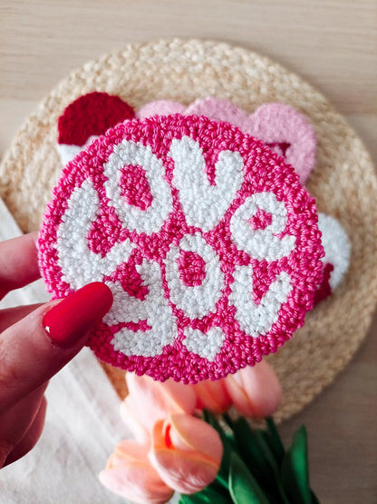 Handmade pink punch needle coaster with white "Love You" lettering, displayed on a woven mat with decorative flowers in the back.