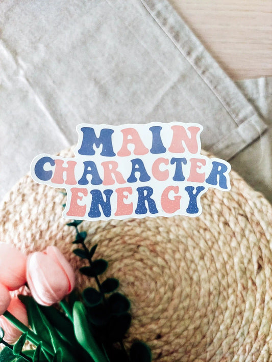 Vinyl sticker with the phrase "Main Character Energy" in blue and pink lettering, photographed on a neutral textured background.