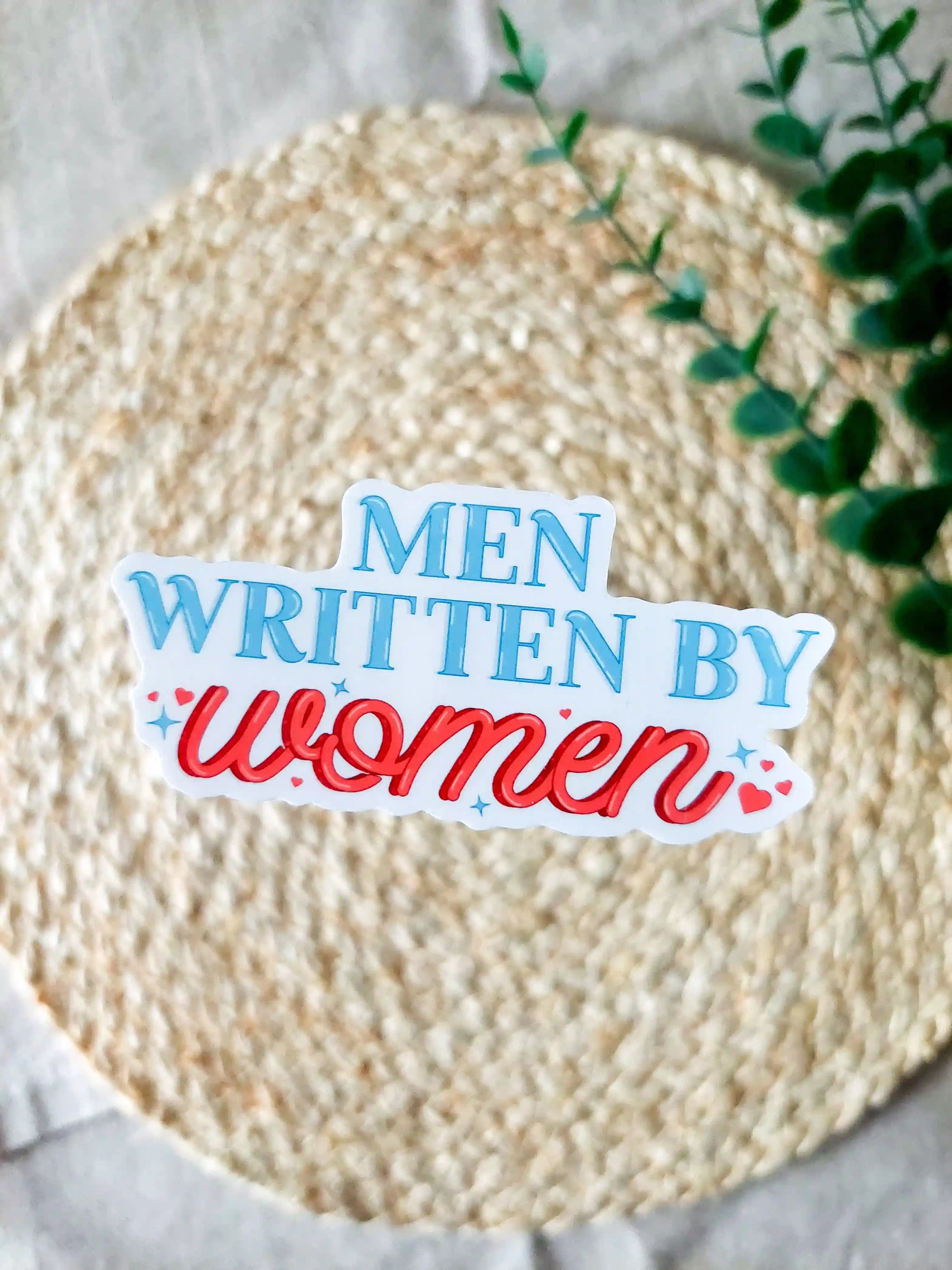 "Men Written By Women" sticker on a rattan placemat, featuring blue and red text with small hearts and stars details.