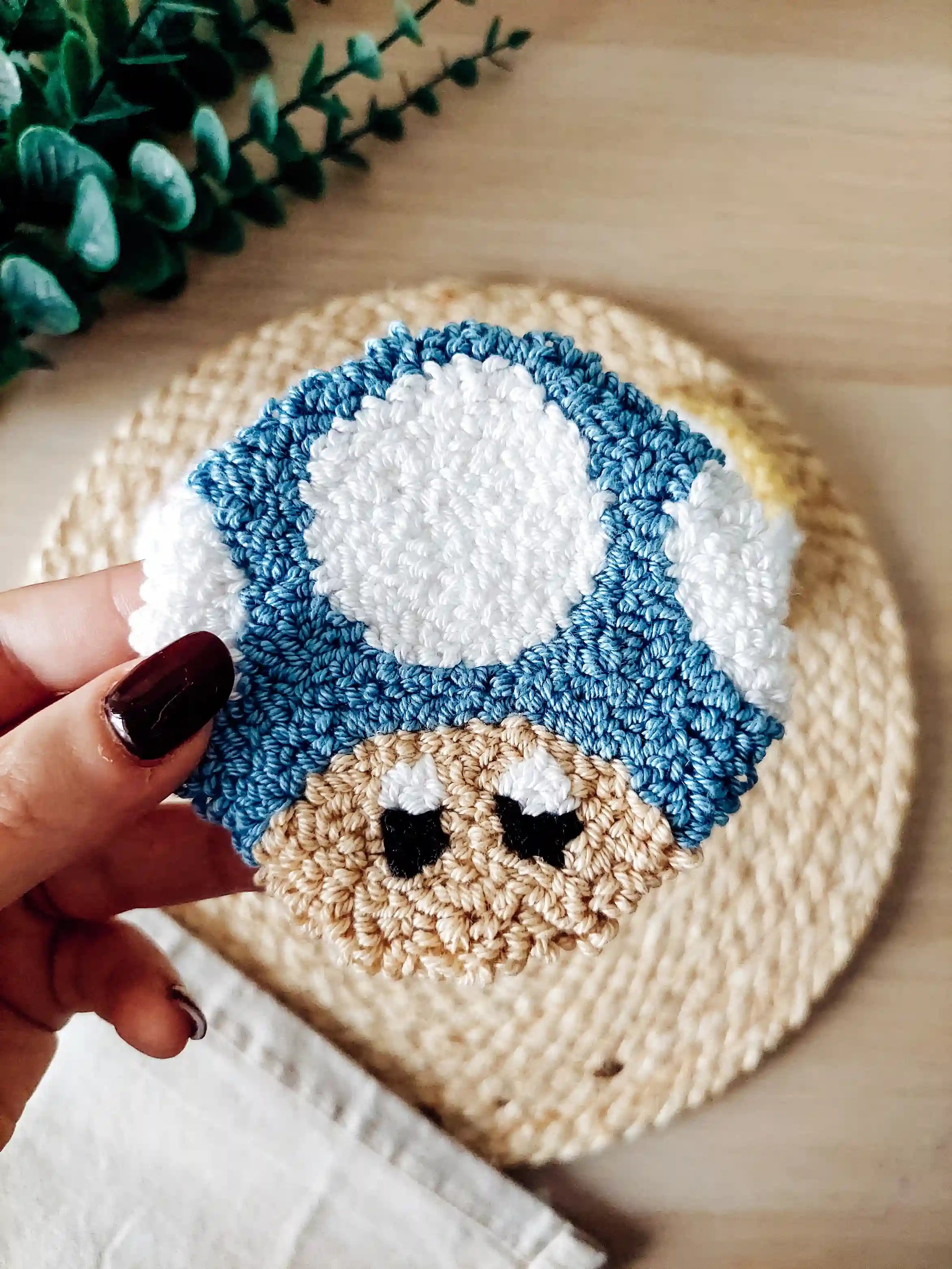 Punch needle mushroom coasters in  blue, inspired by Toad, displayed on a woven mat with decorative flowers.