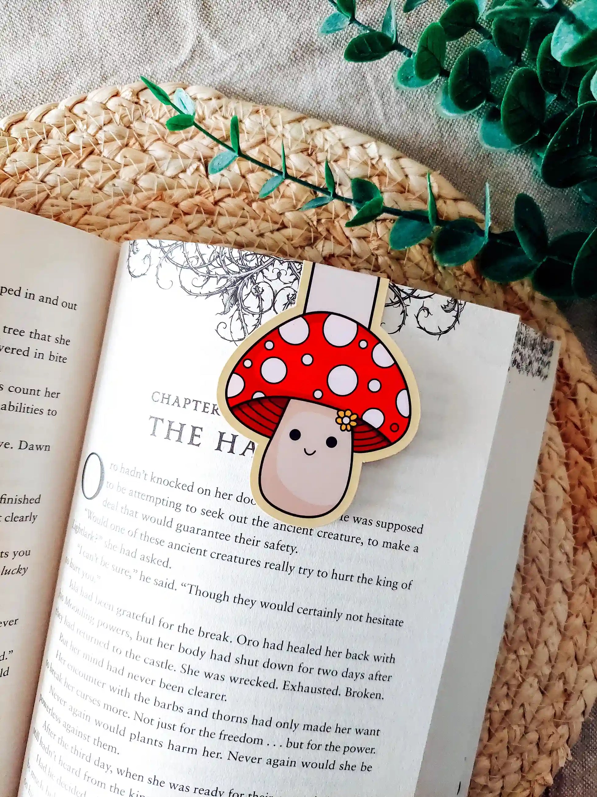 Book with a cute mushroom-shaped magnetic bookmark on an open book, surrounded by green leaves.