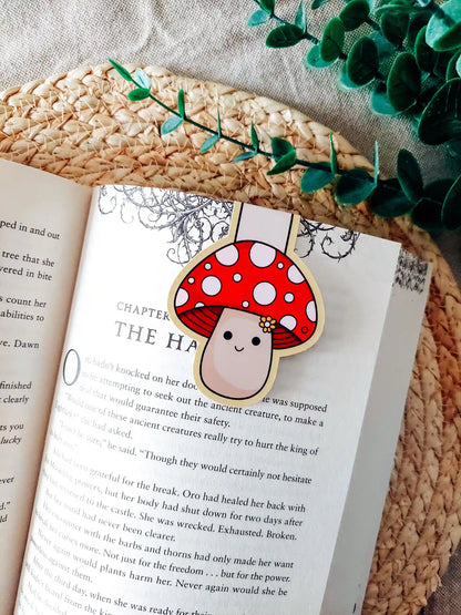 Book with a cute mushroom-shaped magnetic bookmark on an open book, surrounded by green leaves.