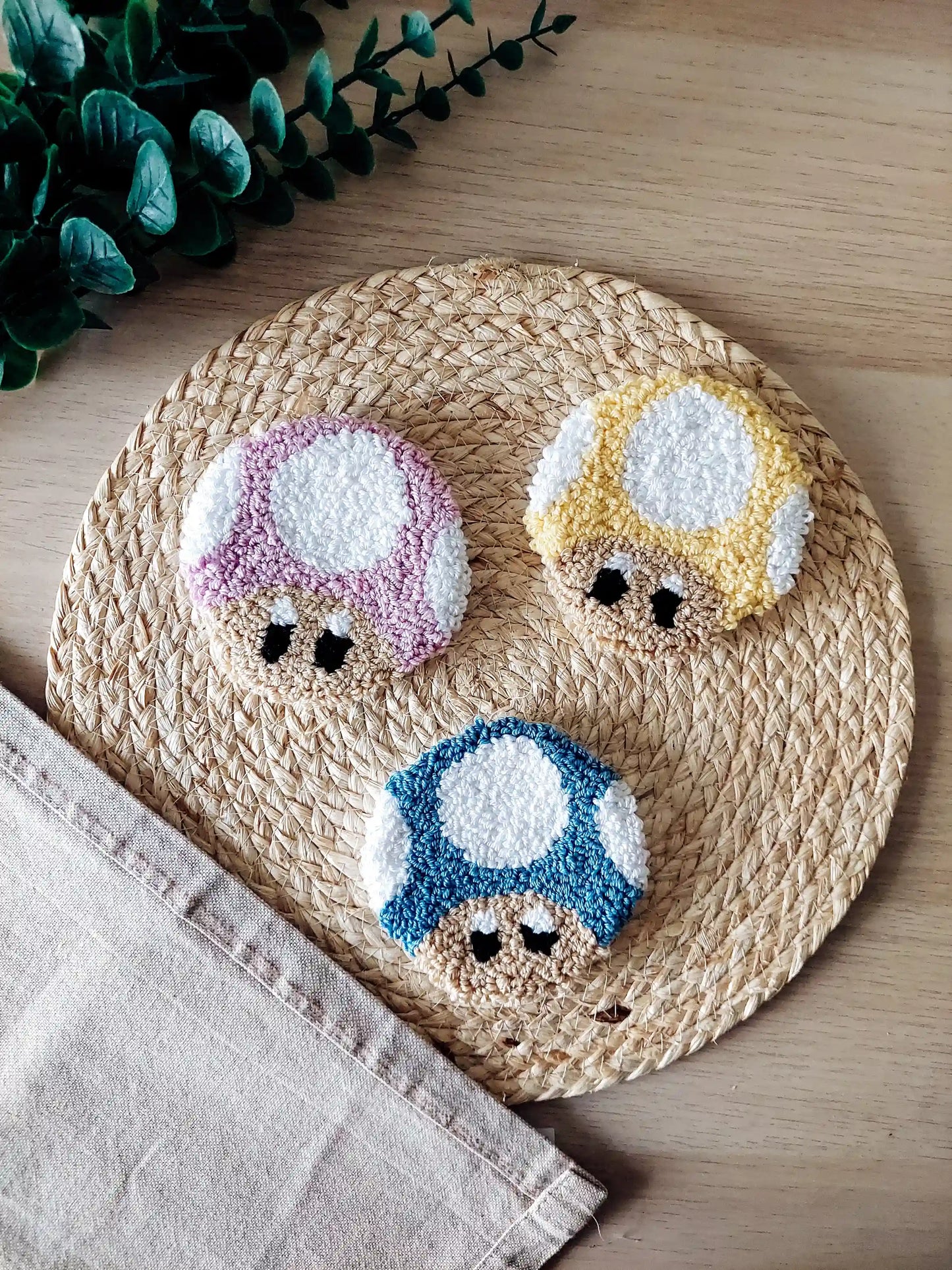 Set of three punch needle mushroom coasters in lilac, yellow and blue, inspired by Toad, displayed on a woven mat with decorative flowers.