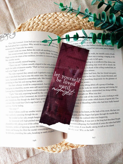 Nightshade bookmark placed on an open book, featuring a dark burgundy background with flying birds, a crossword pattern with book-related words and the text  "Let yourself be loved until night" in white.