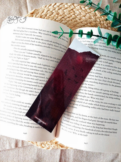 Back of the Nightshade bookmark placed on an open book, featuring a dark burgundy background with flying birds, a crossword pattern with book-related words. 