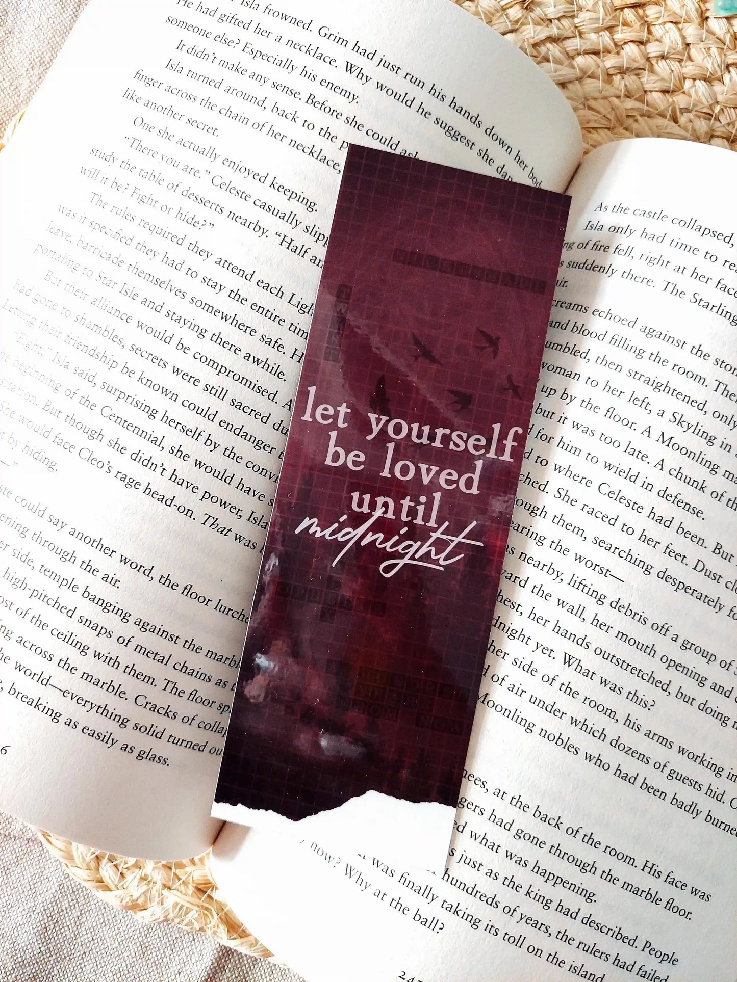 Nightshade bookmark placed on an open book, featuring a dark burgundy background with flying birds, a crossword pattern with book-related words and the text  "Let yourself be loved until night" in white.
