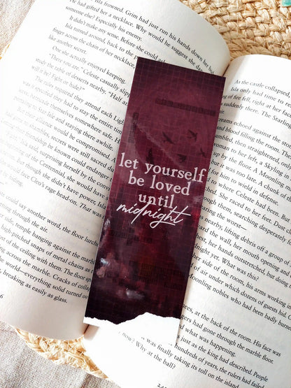 Nightshade bookmark placed on an open book, featuring a dark burgundy background with flying birds, a crossword pattern with book-related words and the text  "Let yourself be loved until night" in white.