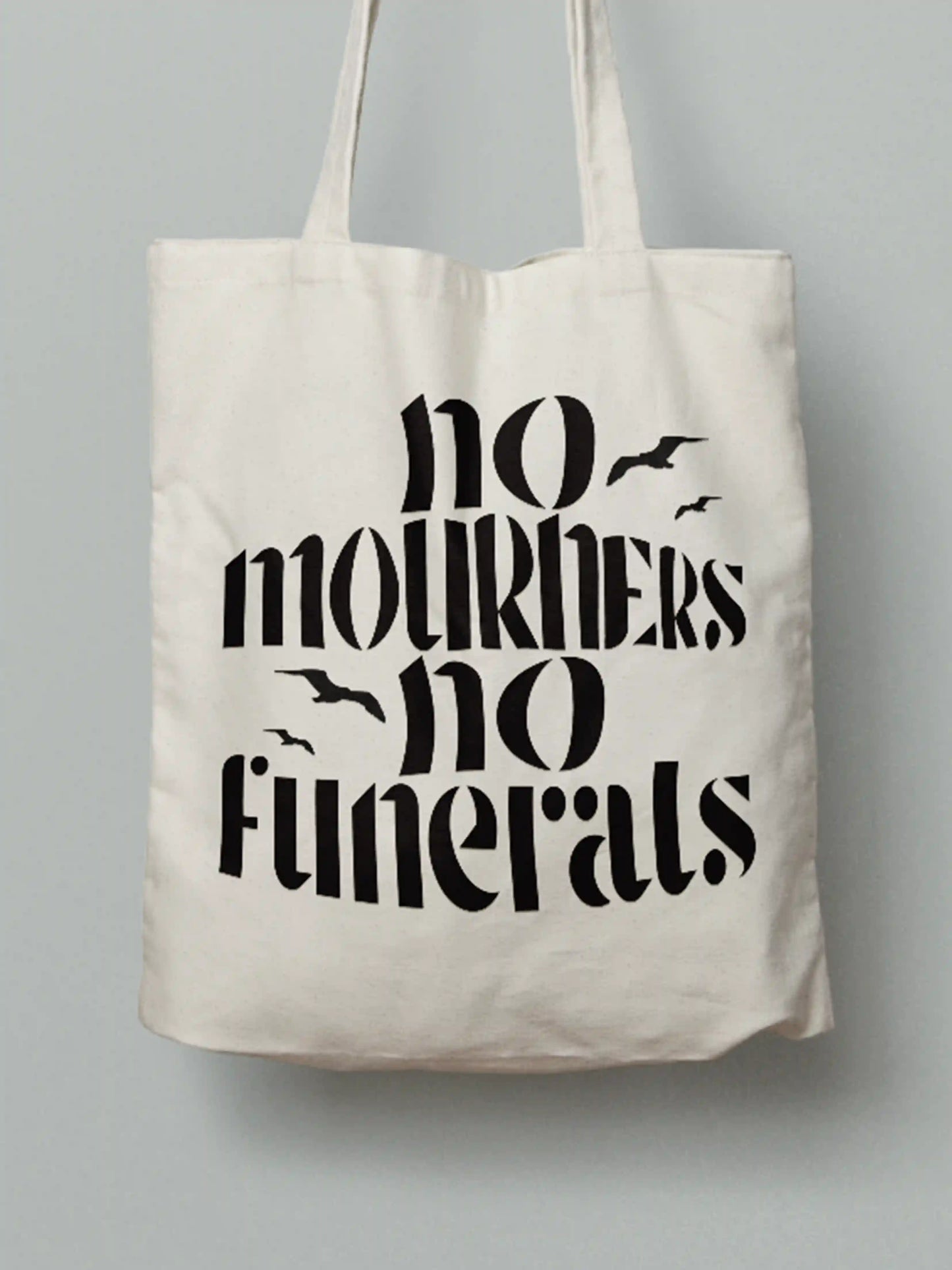 Cotton canvas tote bag with black text "No mourners, no funerals" featuring flying bird silhouettes, photographed against a neutral grey background