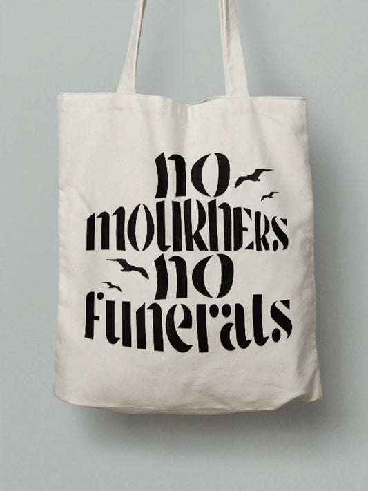 Cotton canvas tote bag with black text "No mourners, no funerals" featuring flying bird silhouettes, photographed against a neutral grey background