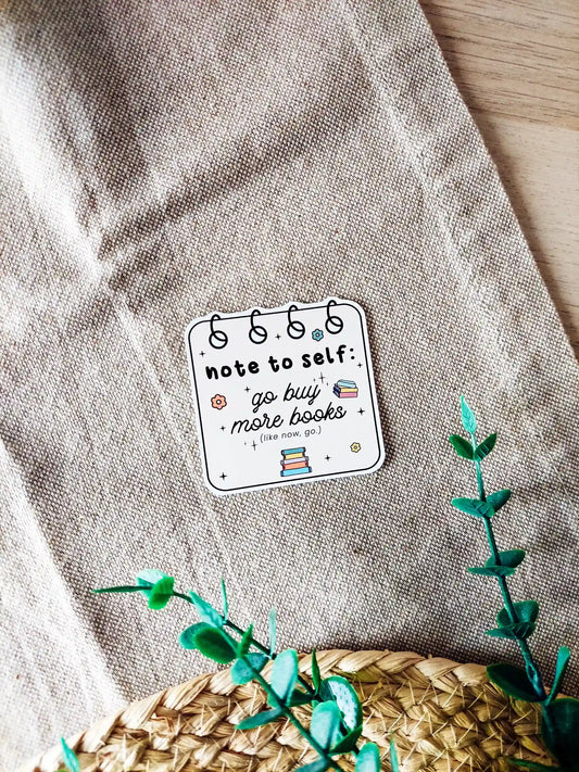 Vinyl sticker of spiral notebook page, with small book illustrations and floral details with the textt "Note to self go buy more books", photographed on a neutral fabric background.