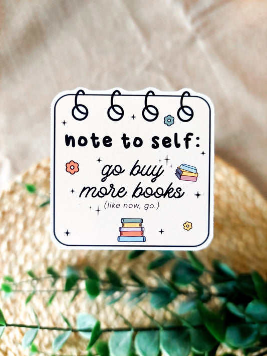 Vinyl sticker of spiral notebook page, with small book illustrations and floral details with the textt "Note to self go buy more books", photographed on a neutral fabric background.