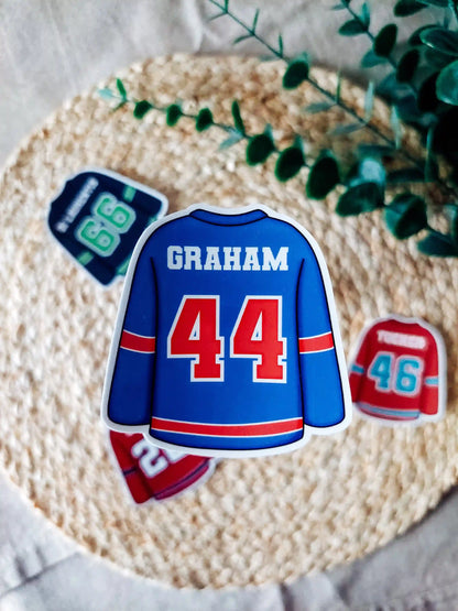 Off Campus inspired blue hockey jersey with "Graham 44" on it, placed on a textured surface with greenery.